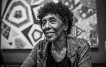 An older woman with curly hair and hoop earrings smiles softly while sitting indoors. She wears a denim jacket over a dark shirt. Abstract geometric art hangs on the wall behind her. The image is in black and white.