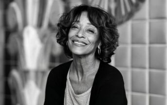 Smiling older woman with curly hair and hoop earrings, wearing a light shirt and dark cardigan, sitting in front of a tiled wall with geometric patterns.