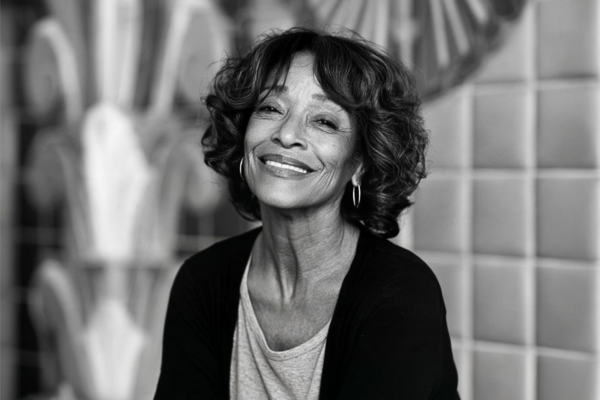 Smiling older woman with curly hair and hoop earrings, wearing a light shirt and dark cardigan, sitting in front of a tiled wall with geometric patterns.