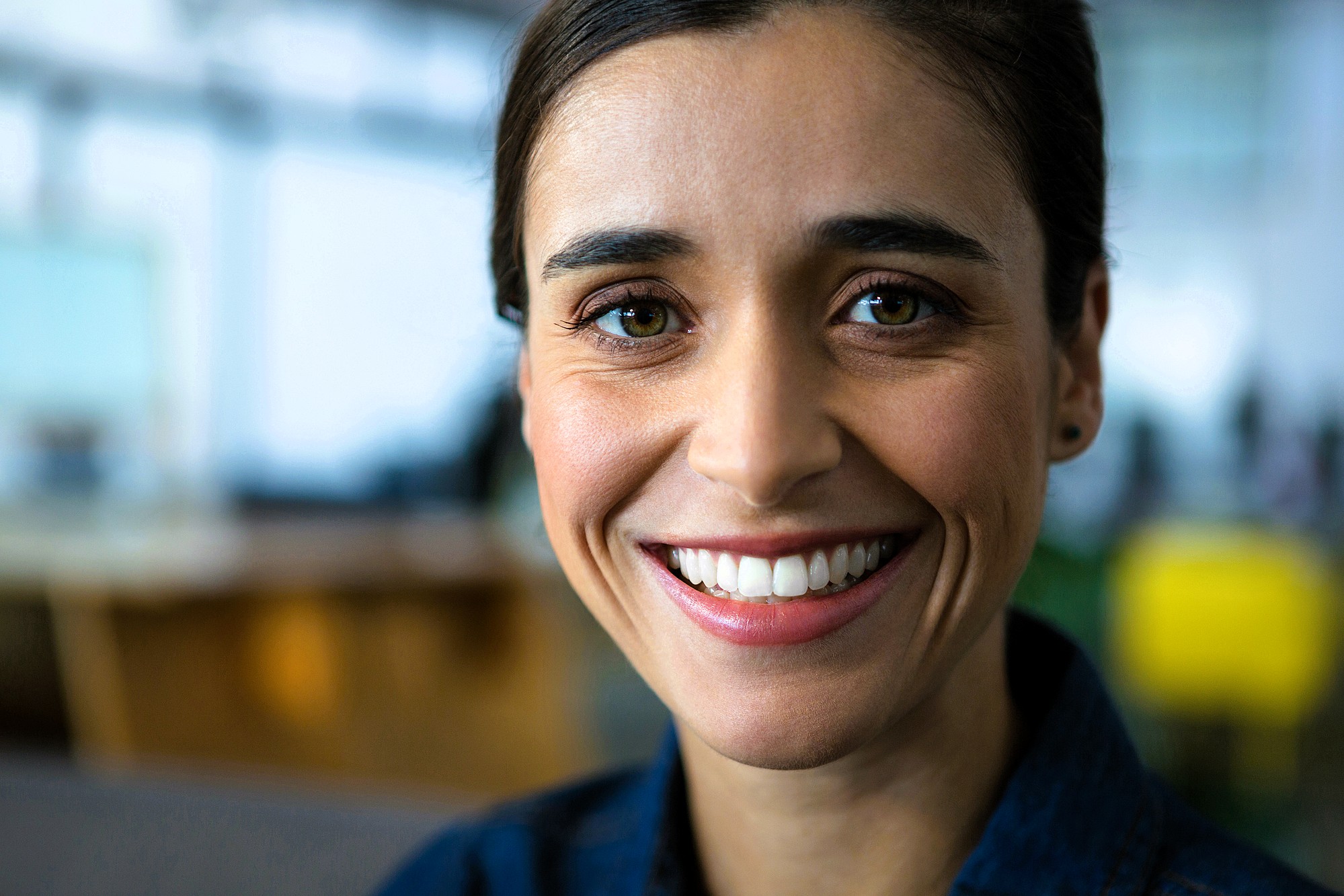 A person with dark hair smiles warmly, wearing a denim shirt. The background is softly blurred, suggesting an indoor setting. The lighting highlights their face, giving the image a cheerful and welcoming vibe.