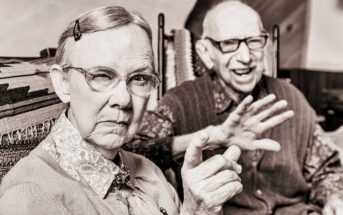 An elderly woman with glasses and a hairpin gestures with a finger while a smiling elderly man with glasses and a sweater vest sits beside her, gesturing with his hand. They are indoors, seated on patterned chairs. The image is in sepia tone.