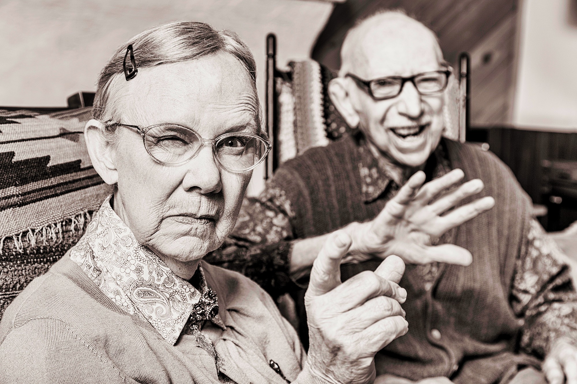 An elderly woman with glasses and a hairpin gestures with a finger while a smiling elderly man with glasses and a sweater vest sits beside her, gesturing with his hand. They are indoors, seated on patterned chairs. The image is in sepia tone.