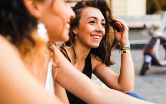 Two women sitting outdoors, smiling and engaging in conversation. One woman, with long hair, is in focus wearing a black top and jewelry, while the other, wearing a white top, is blurred in the foreground. A scooter is in the background.
