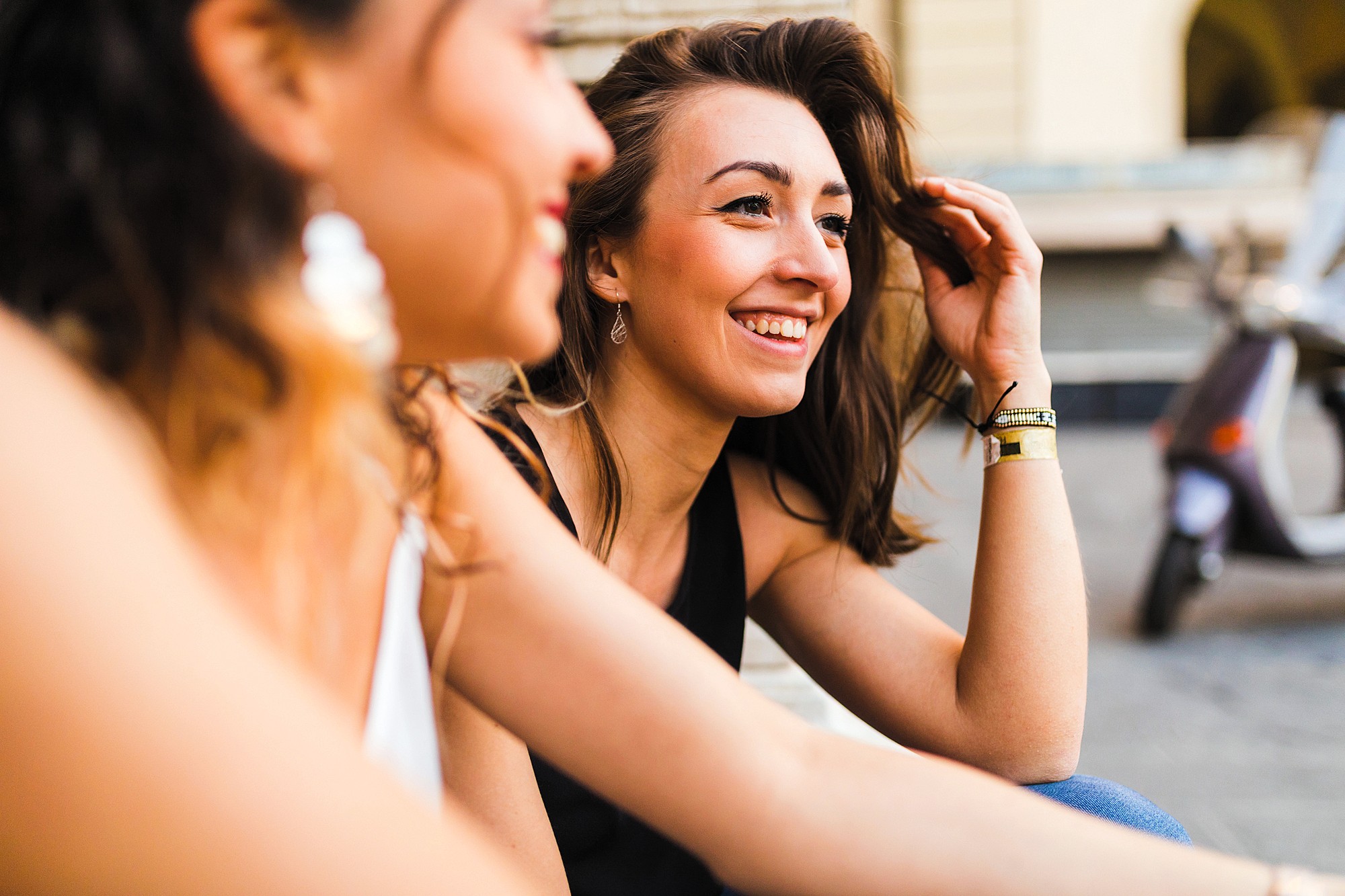 Two women sitting outdoors, smiling and engaging in conversation. One woman, with long hair, is in focus wearing a black top and jewelry, while the other, wearing a white top, is blurred in the foreground. A scooter is in the background.