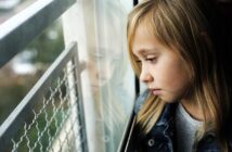 A young girl with long blonde hair looks out a window, her reflection visible in the glass. She wears a denim jacket and appears thoughtful or pensive as she gazes outside. The background is blurred.