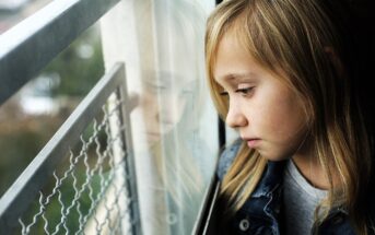 A young girl with long blonde hair looks out a window, her reflection visible in the glass. She wears a denim jacket and appears thoughtful or pensive as she gazes outside. The background is blurred.