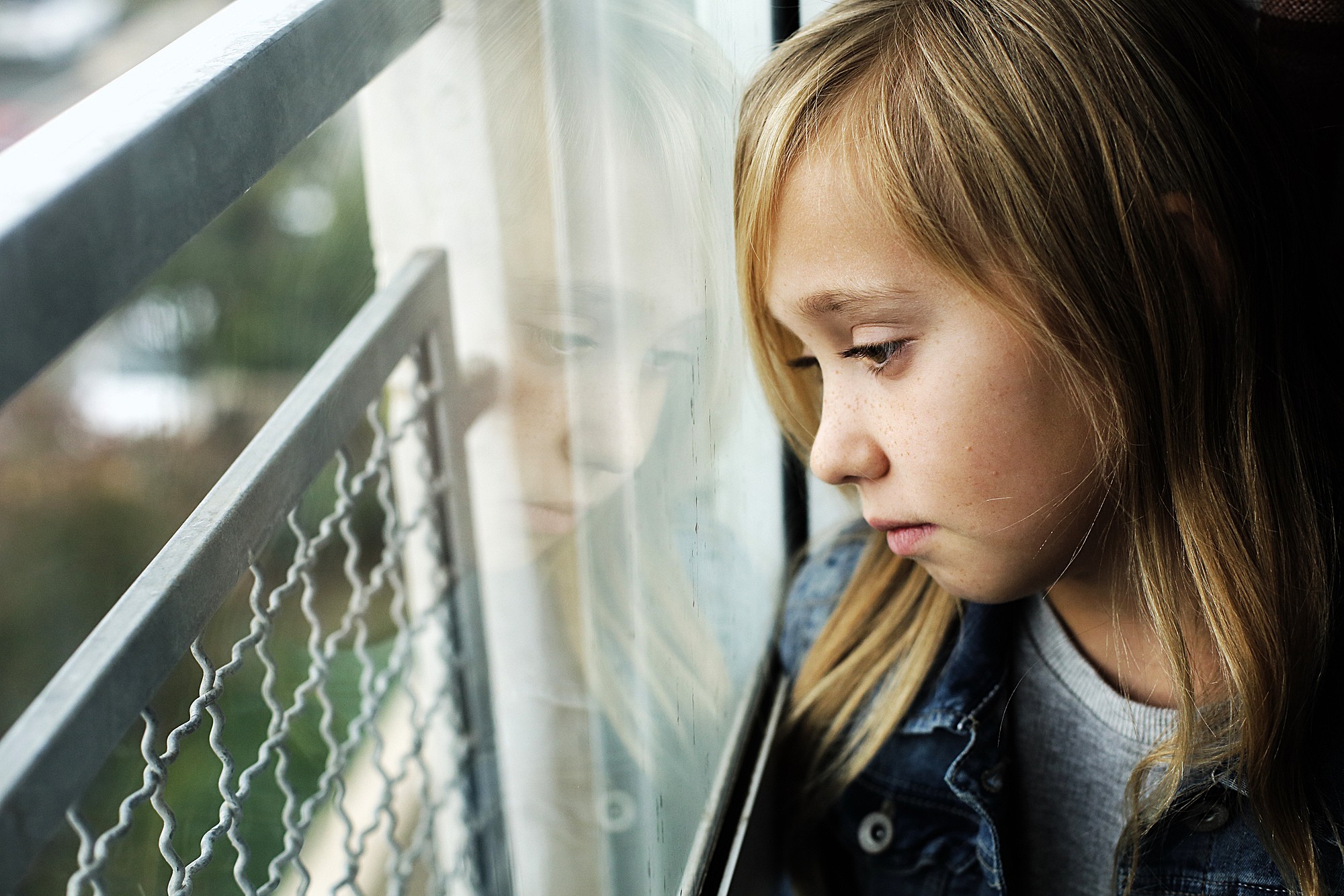 A young girl with long blonde hair looks out a window, her reflection visible in the glass. She wears a denim jacket and appears thoughtful or pensive as she gazes outside. The background is blurred.