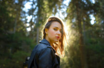 A woman with shoulder-length hair stands in a sunlit forest, wearing a black leather jacket. She looks back over her shoulder, with sunlight creating a halo effect around her hair against the green, blurry background.