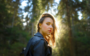A woman with shoulder-length hair stands in a sunlit forest, wearing a black leather jacket. She looks back over her shoulder, with sunlight creating a halo effect around her hair against the green, blurry background.