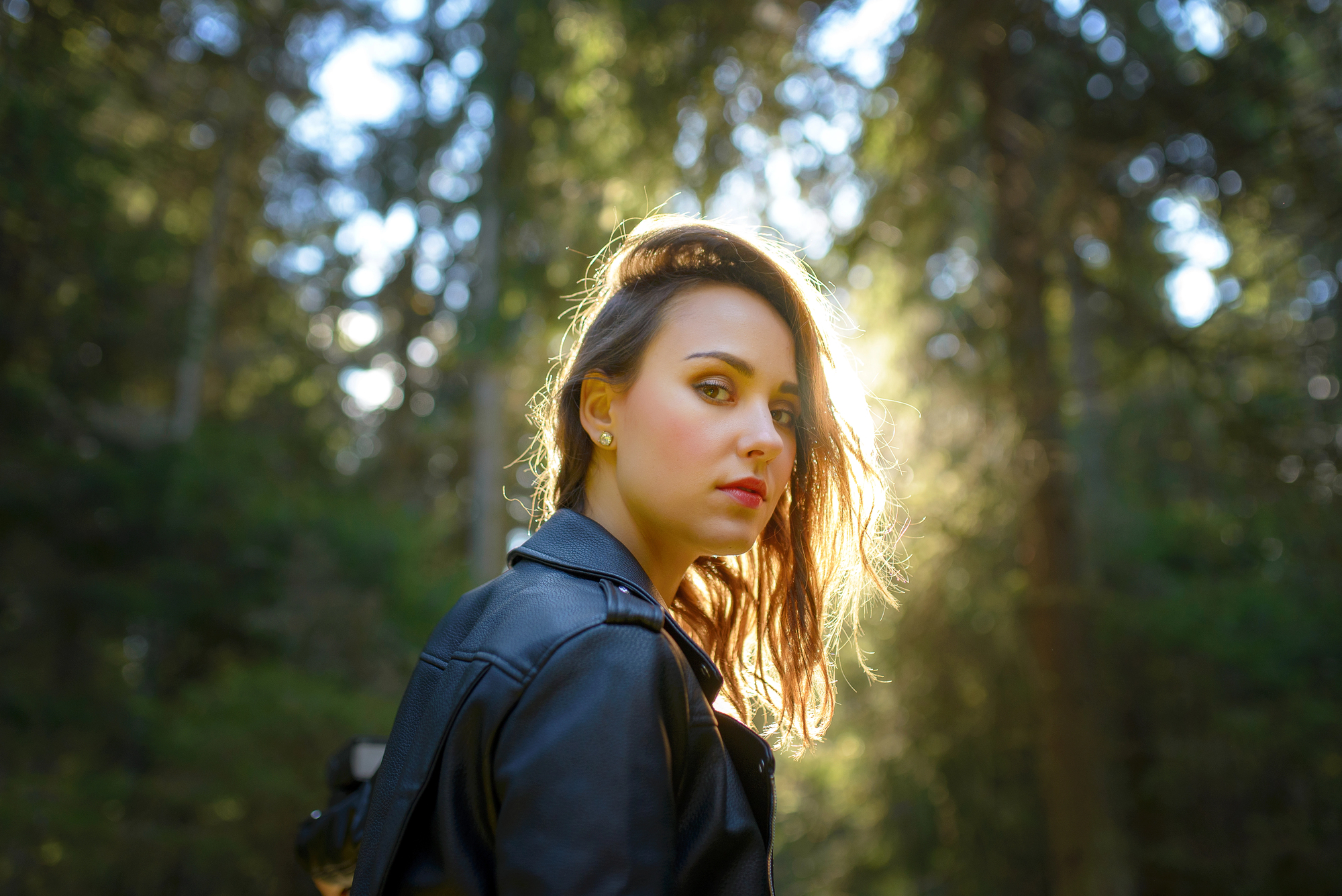 A woman with shoulder-length hair stands in a sunlit forest, wearing a black leather jacket. She looks back over her shoulder, with sunlight creating a halo effect around her hair against the green, blurry background.