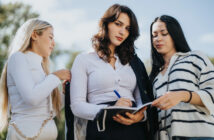 Three women stand outdoors. The woman in the middle writes in a notebook, wearing a white blouse with a dark jacket draped over her shoulder. The women on either side observe, with a bright, clear sky and trees in the background.