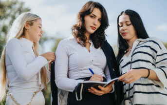 Three women stand outdoors. The woman in the middle writes in a notebook, wearing a white blouse with a dark jacket draped over her shoulder. The women on either side observe, with a bright, clear sky and trees in the background.