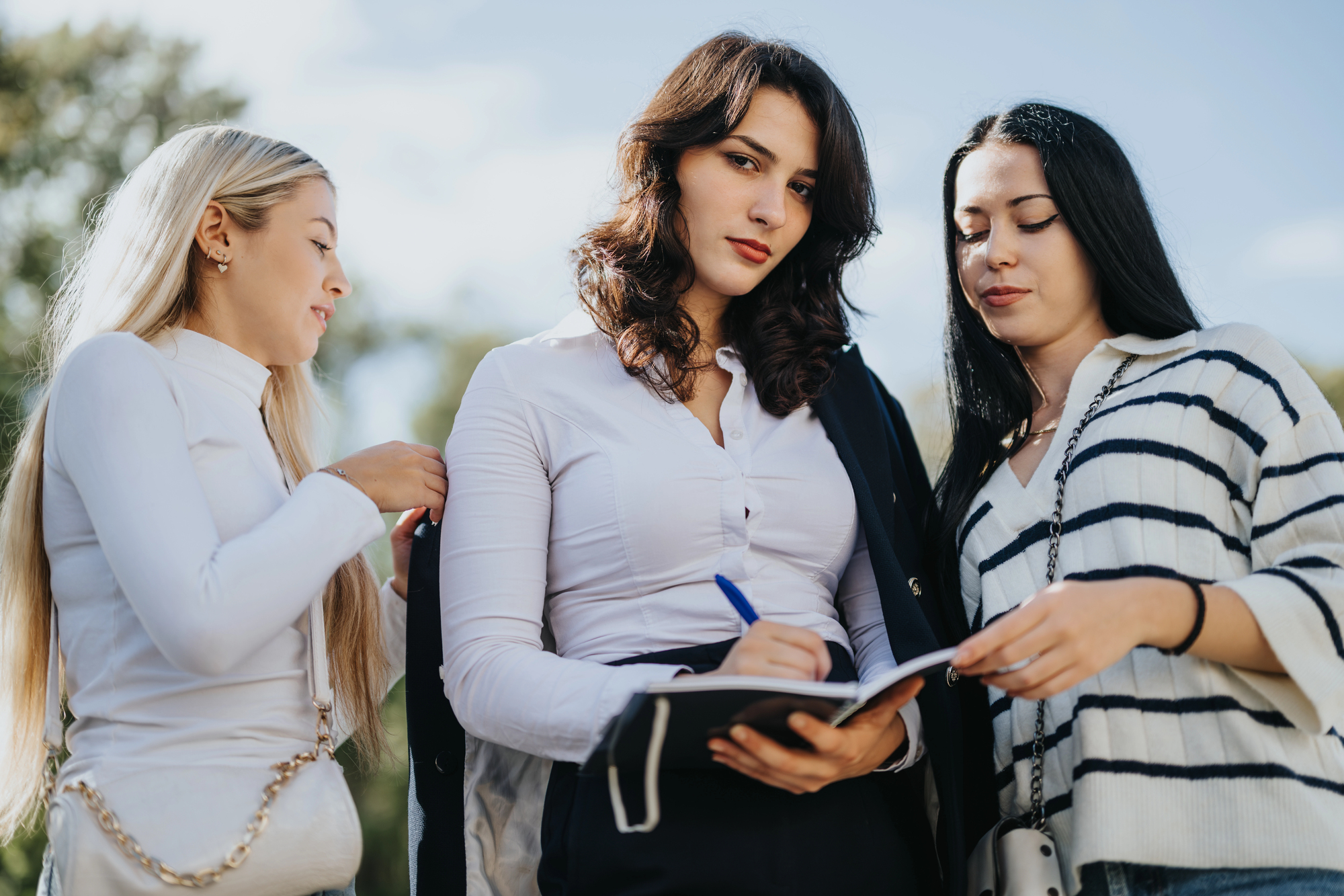 Three women stand outdoors. The woman in the middle writes in a notebook, wearing a white blouse with a dark jacket draped over her shoulder. The women on either side observe, with a bright, clear sky and trees in the background.