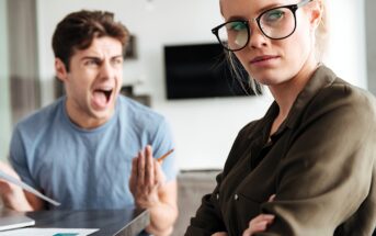 A woman with glasses stands with her arms crossed and an annoyed expression, looking away. A man in the background wears a blue shirt, holding papers and shouting, appearing frustrated. The setting seems to be an indoor office space.