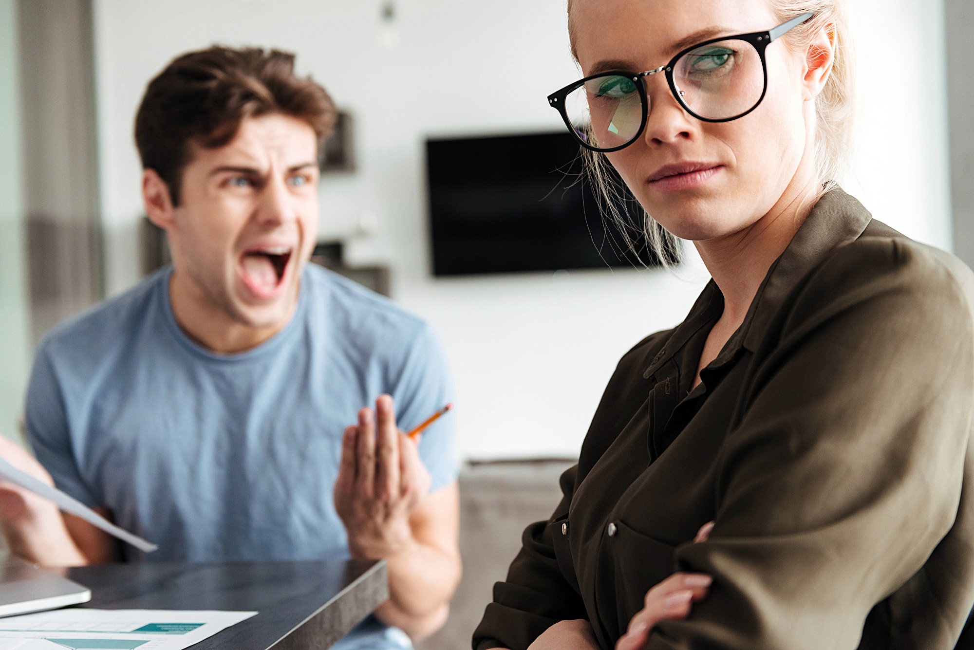 A woman with glasses stands with her arms crossed and an annoyed expression, looking away. A man in the background wears a blue shirt, holding papers and shouting, appearing frustrated. The setting seems to be an indoor office space.