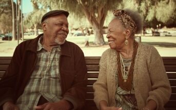 An elderly man and woman sit on a park bench, smiling warmly at each other. They are dressed in casual, comfortable clothing, with trees and soft sunlight in the background.