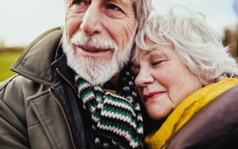 Elderly couple embracing outdoors. The man wears a striped scarf and jacket, while the woman has her eyes closed and leans on his shoulder, wrapped in a yellow scarf. They appear content and peaceful, with a blurry landscape in the background.
