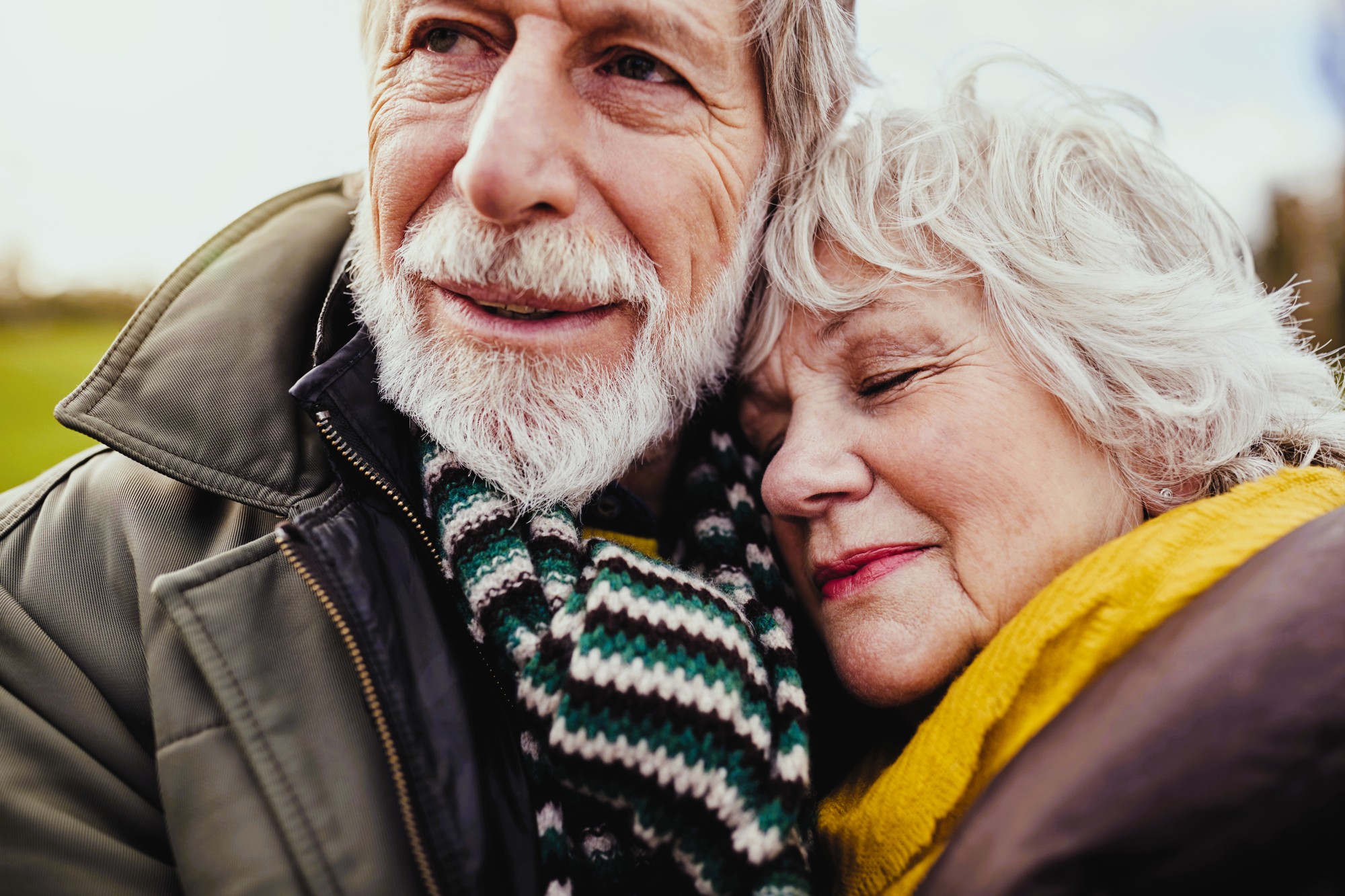 Elderly couple embracing outdoors. The man wears a striped scarf and jacket, while the woman has her eyes closed and leans on his shoulder, wrapped in a yellow scarf. They appear content and peaceful, with a blurry landscape in the background.
