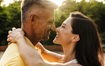 A couple embraces and smiles warmly at each other outdoors, with a blurred background of green trees and sunlight. The woman has long dark hair and the man has short hair and a beard, both appear happy and close.
