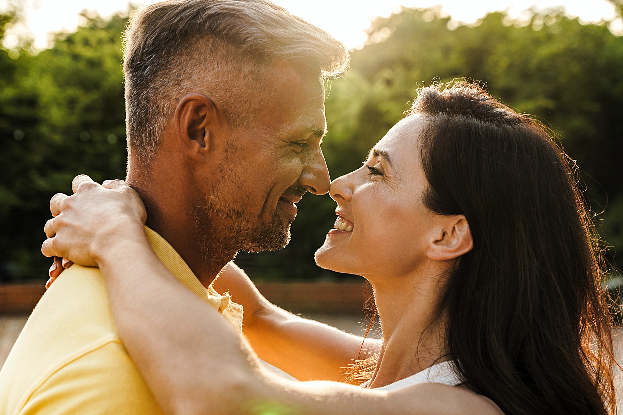 A couple embraces and smiles warmly at each other outdoors, with a blurred background of green trees and sunlight. The woman has long dark hair and the man has short hair and a beard, both appear happy and close.
