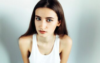 A woman with long brown hair and green eyes is looking directly at the camera. She is wearing a sleeveless white top and is set against a plain background. The lighting highlights her facial features and expression.