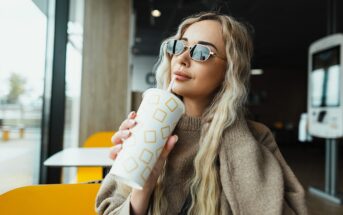 A woman with long blonde hair and sunglasses holds a disposable cup with a geometric design. She is sitting indoors by a large window, wearing a brown sweater, with a contemplative expression. The setting appears casual and modern.