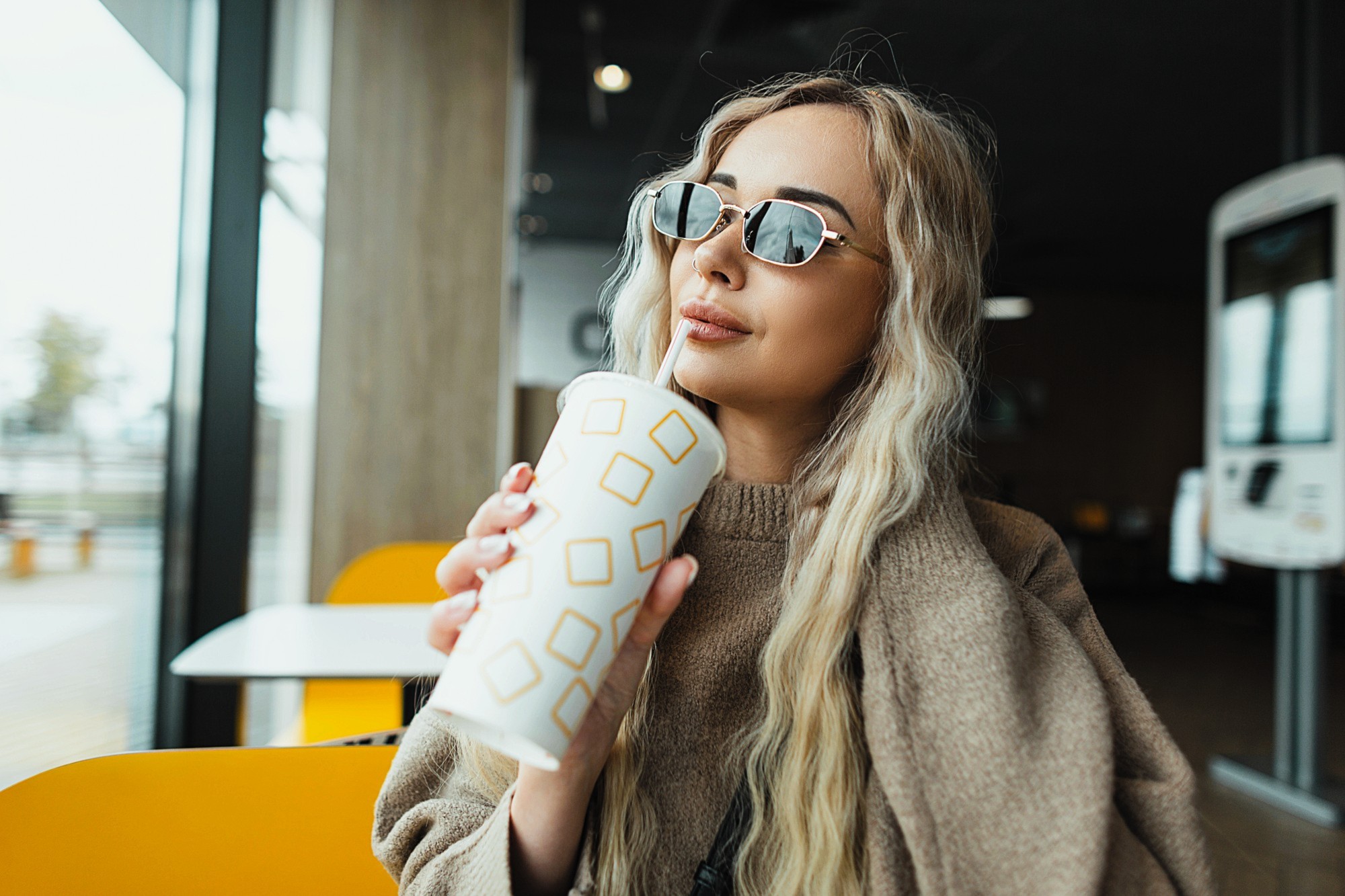 A woman with long blonde hair and sunglasses holds a disposable cup with a geometric design. She is sitting indoors by a large window, wearing a brown sweater, with a contemplative expression. The setting appears casual and modern.