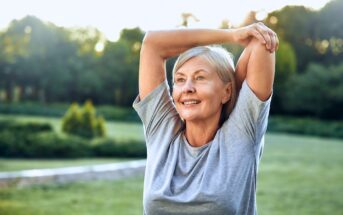 Elderly woman with gray hair smiling while stretching her arms overhead in a green park, wearing a gray T-shirt. The background features lush trees and a clear sky, suggesting a serene outdoor setting.