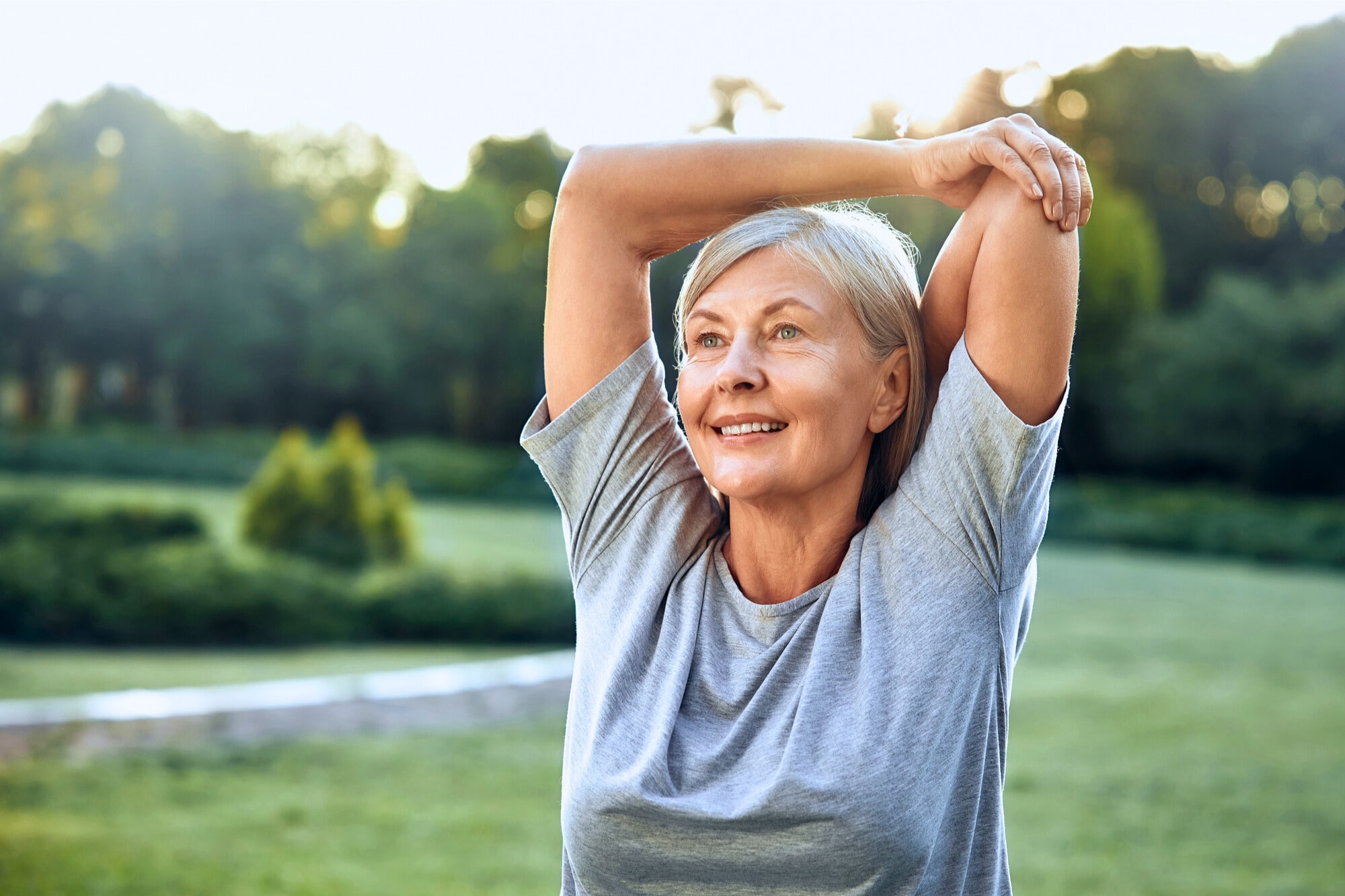 Elderly woman with gray hair smiling while stretching her arms overhead in a green park, wearing a gray T-shirt. The background features lush trees and a clear sky, suggesting a serene outdoor setting.