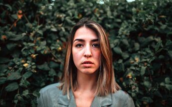 A person with shoulder-length hair stands in front of dense green foliage, wearing a gray plaid jacket. They have a neutral expression and are centered in the image, with the leaves providing a textured background.