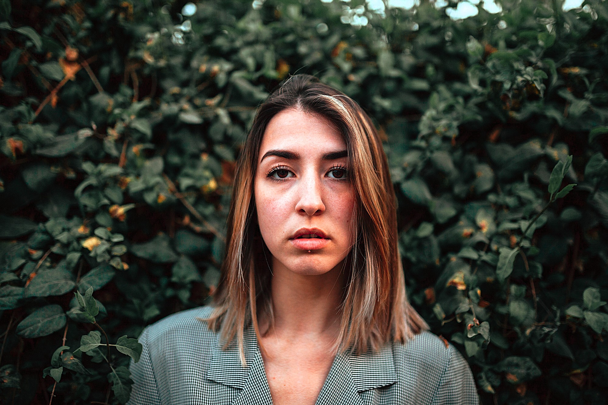 A person with shoulder-length hair stands in front of dense green foliage, wearing a gray plaid jacket. They have a neutral expression and are centered in the image, with the leaves providing a textured background.