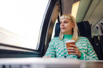 A person with blonde hair and a green patterned sweater is sitting on a train near a window, holding a disposable coffee cup and looking outside. The interior of the train is modern and well-lit.