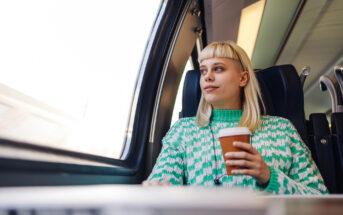 A person with blonde hair and a green patterned sweater is sitting on a train near a window, holding a disposable coffee cup and looking outside. The interior of the train is modern and well-lit.