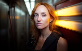 A woman with long red hair stands in a narrow hallway, illuminated by a warm yellow light on the wall, looking directly at the camera with a calm expression.