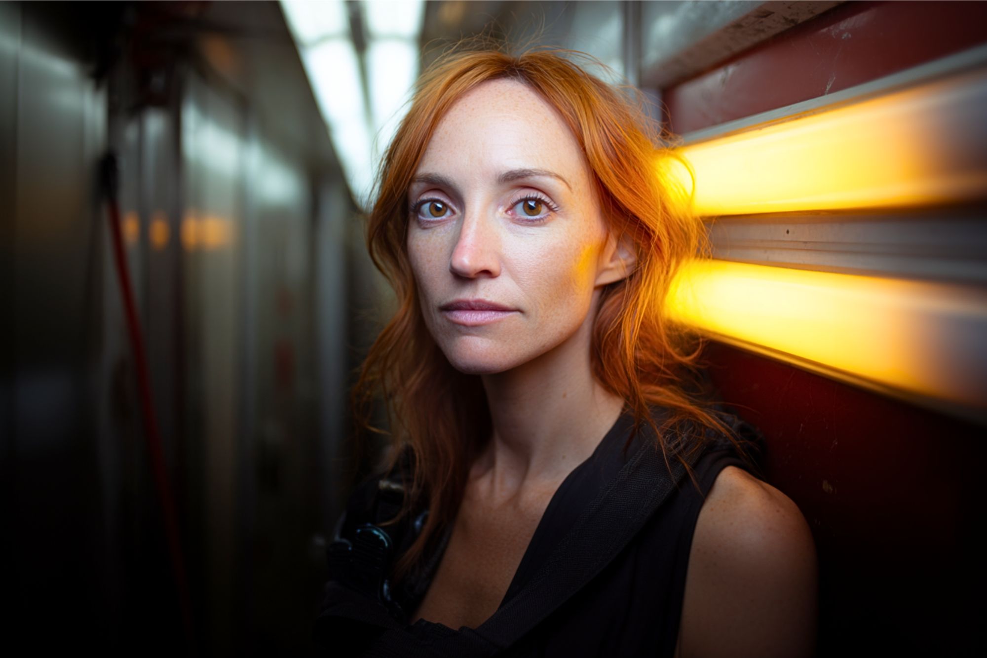 A woman with long red hair stands in a narrow hallway, illuminated by a warm yellow light on the wall, looking directly at the camera with a calm expression.