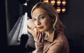 A woman with blonde hair and light eyes leans on a mirror lit with bright bulbs, gazing at the camera. She wears a beige jacket and has colorful manicured nails. Warm lights glow in the background.
