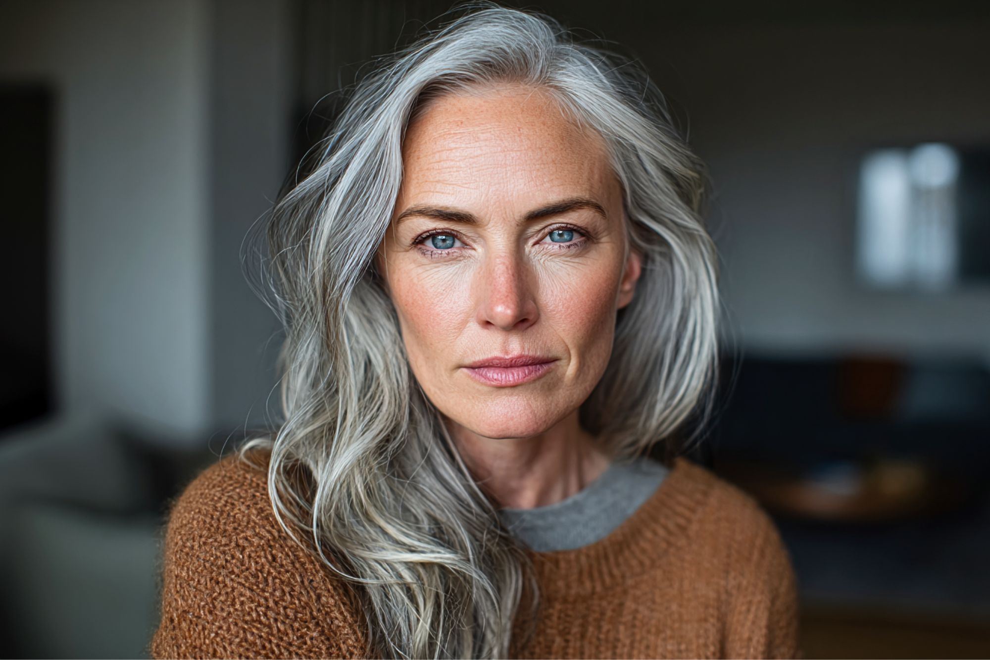 A woman with long gray hair and blue eyes wears a brown sweater, gazing directly at the camera with a calm, confident expression. The background is softly blurred.