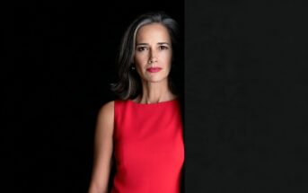 A woman with shoulder-length gray hair wearing a red sleeveless dress, partially obscured by a black panel on the right side of the frame.