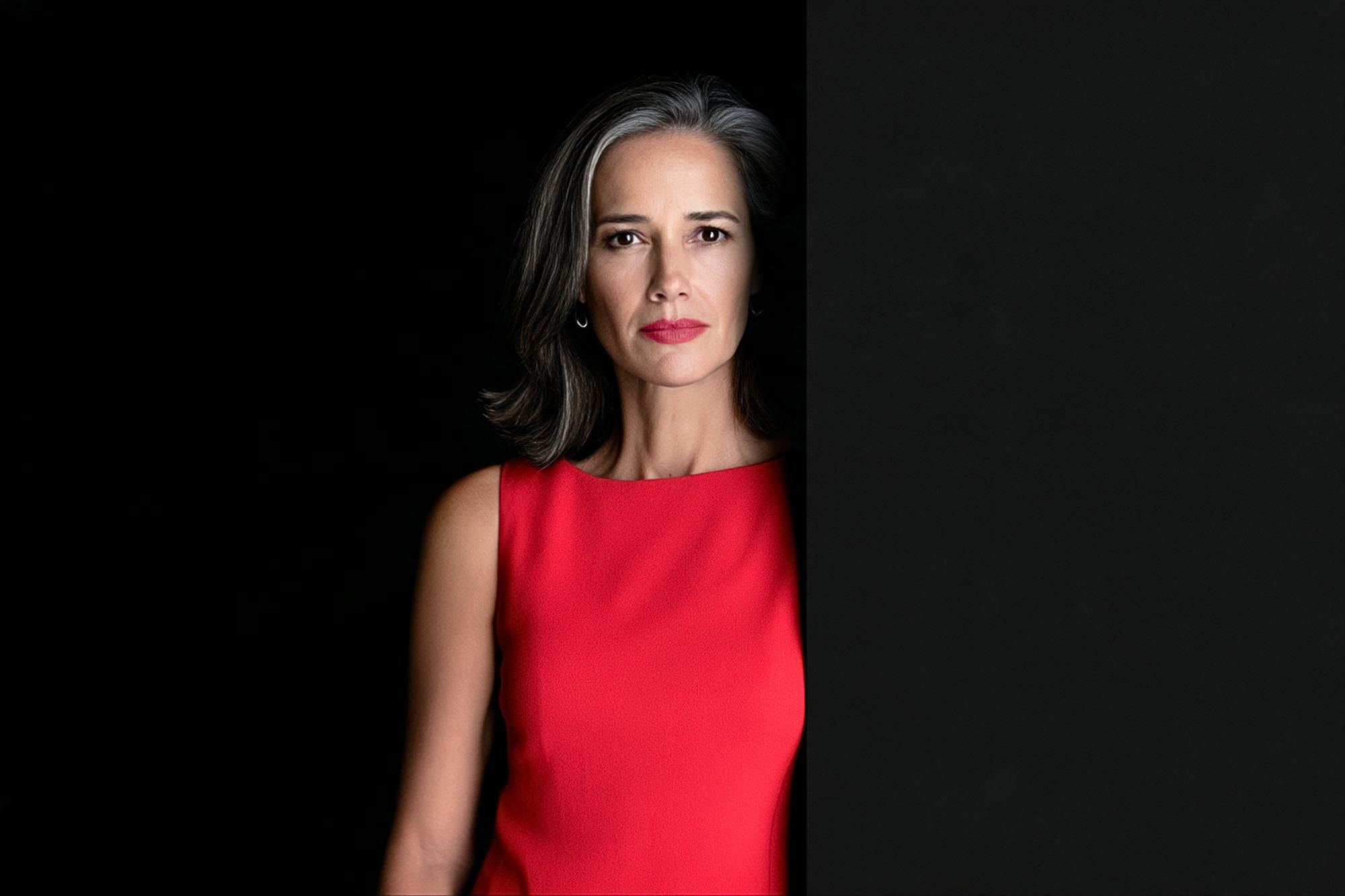 A woman with shoulder-length gray hair wearing a red sleeveless dress, partially obscured by a black panel on the right side of the frame.