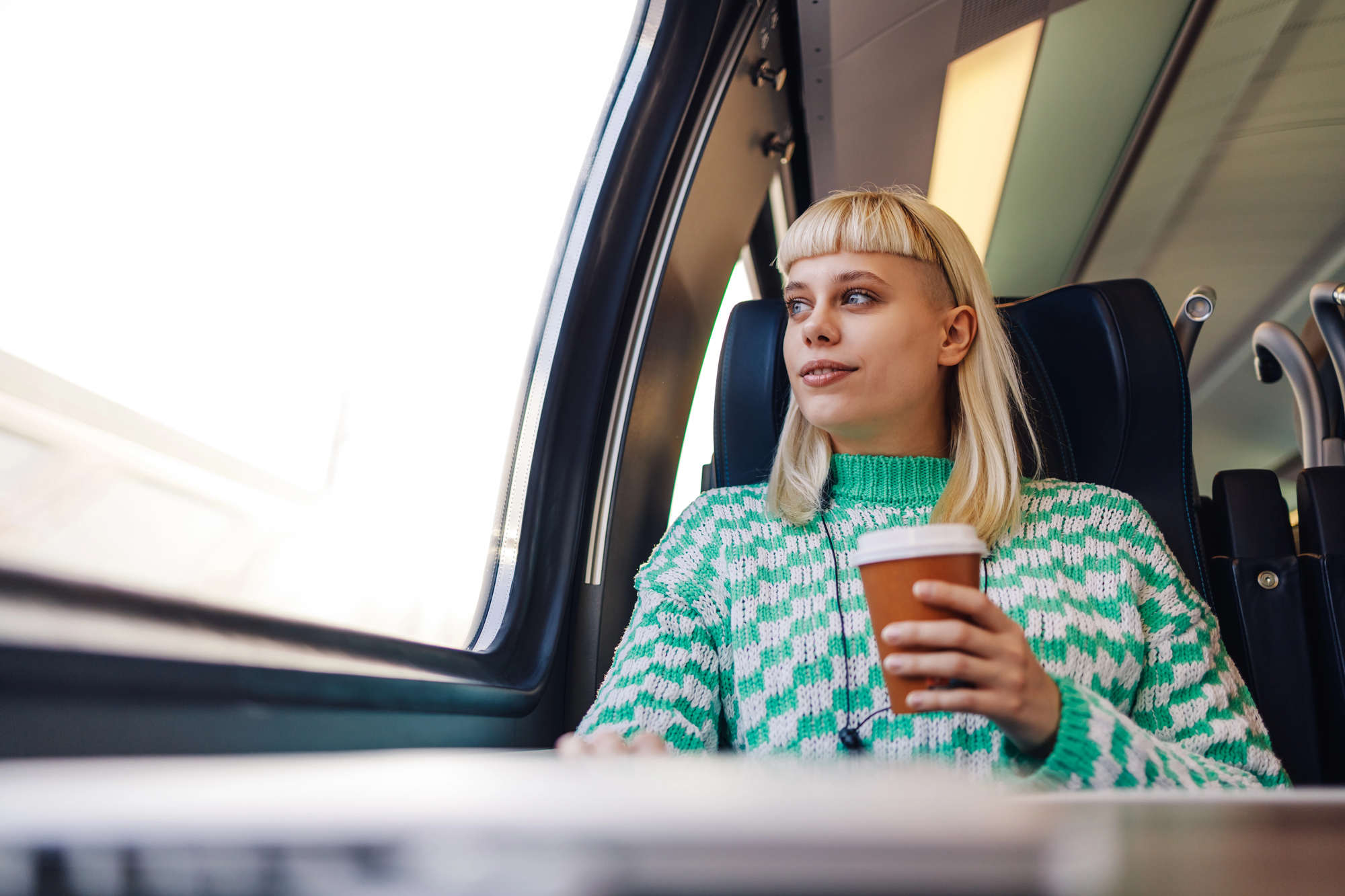 A person with blonde hair and a green patterned sweater is sitting on a train near a window, holding a disposable coffee cup and looking outside. The interior of the train is modern and well-lit.
