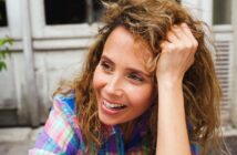 A person with curly hair smiles while leaning their head on their hand. They are wearing a colorful plaid shirt and appear to be outdoors in a casual setting.