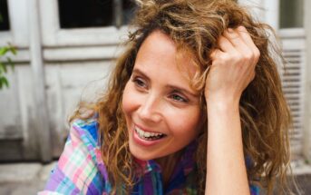 A person with curly hair smiles while leaning their head on their hand. They are wearing a colorful plaid shirt and appear to be outdoors in a casual setting.