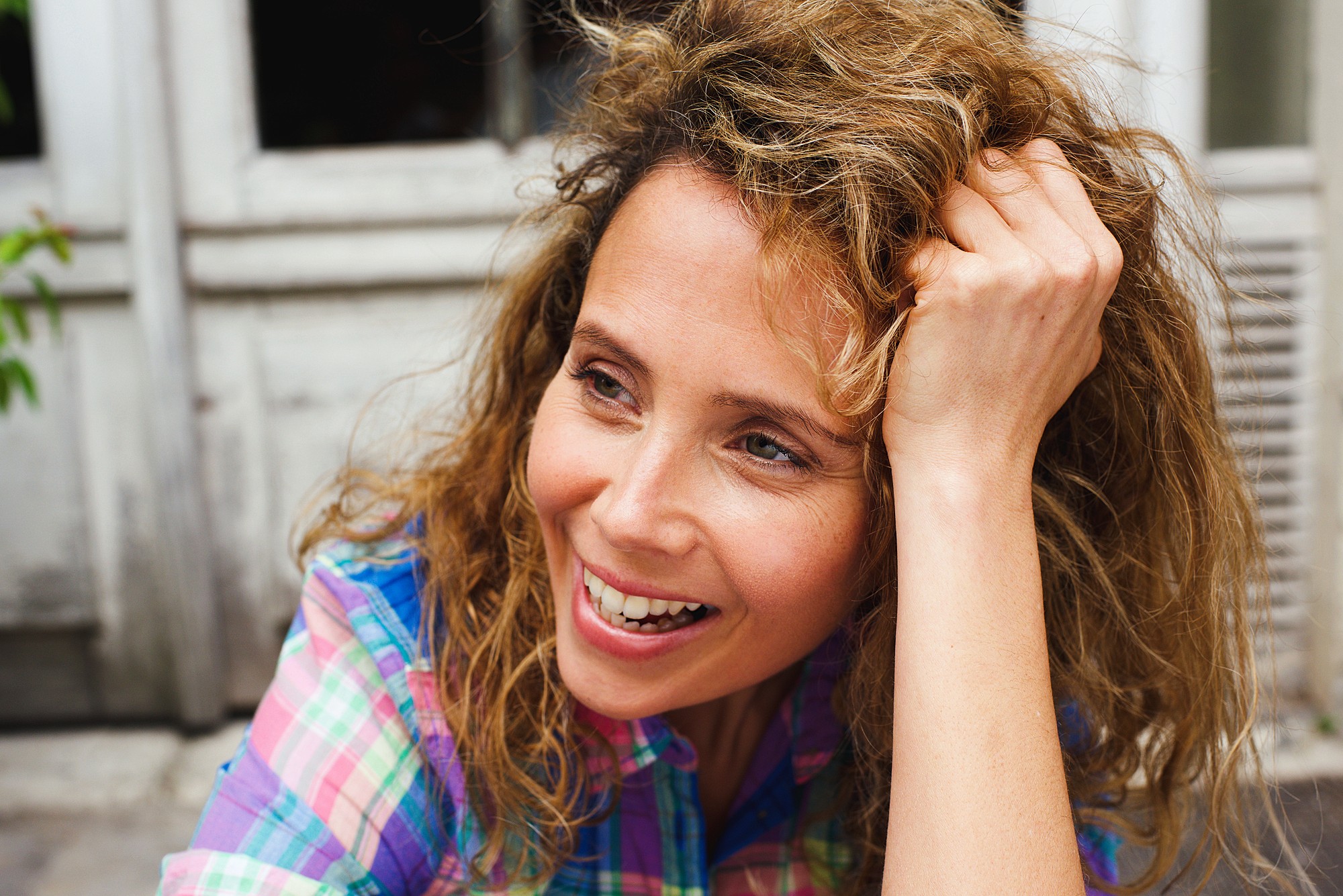 A person with curly hair smiles while leaning their head on their hand. They are wearing a colorful plaid shirt and appear to be outdoors in a casual setting.