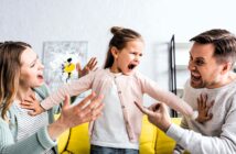 A young girl stands on a yellow sofa, yelling with arms outstretched. A man and woman sit on either side, gesturing animatedly and looking at her. The background has a shelf and abstract painting.