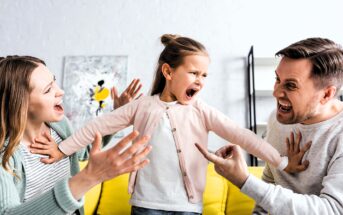 A young girl stands on a yellow sofa, yelling with arms outstretched. A man and woman sit on either side, gesturing animatedly and looking at her. The background has a shelf and abstract painting.
