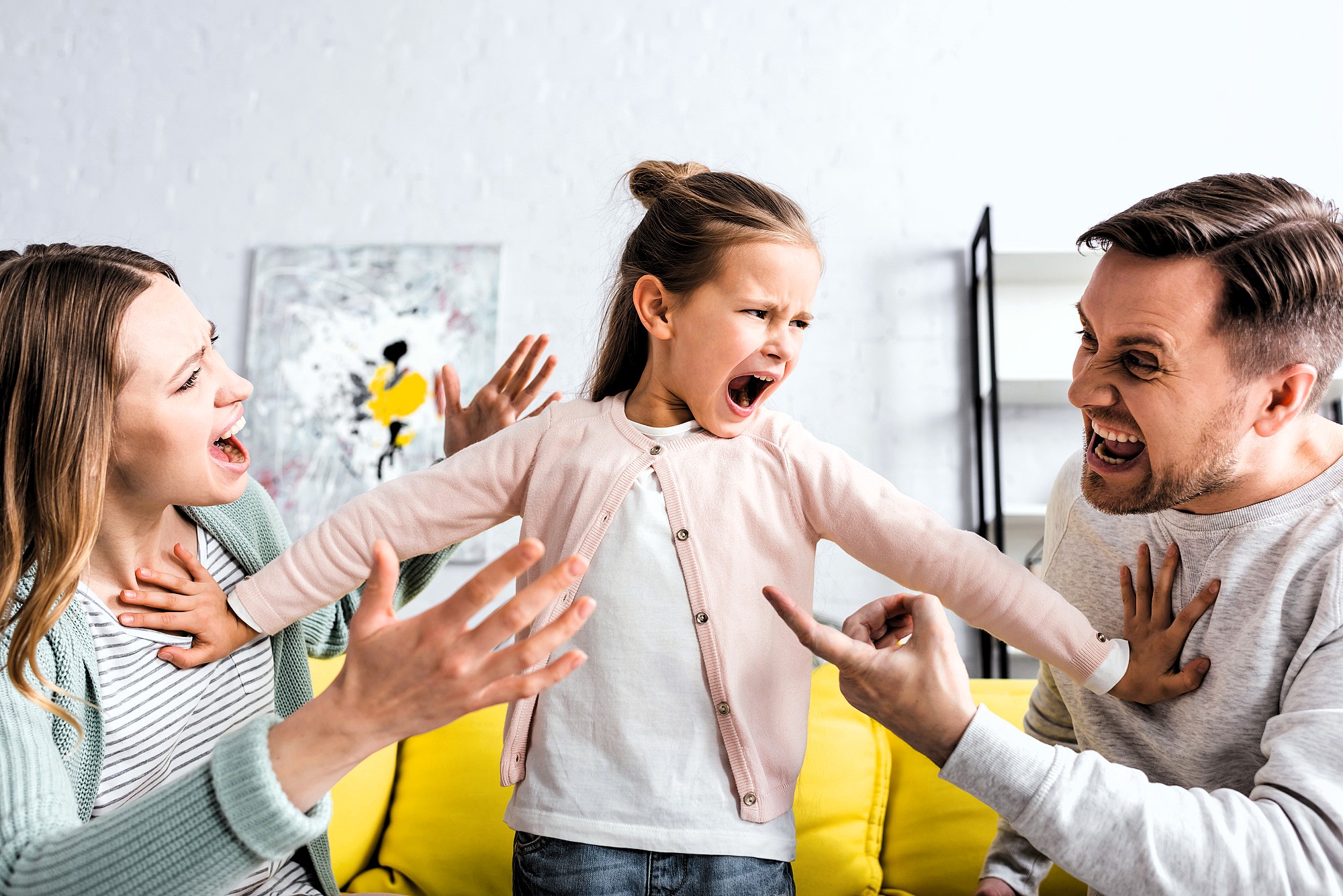 A young girl stands on a yellow sofa, yelling with arms outstretched. A man and woman sit on either side, gesturing animatedly and looking at her. The background has a shelf and abstract painting.