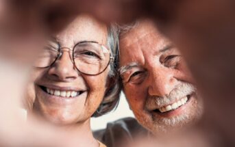 A close-up of a smiling elderly couple. Both are wearing glasses and have gray hair. They are framed by two hands forming a heart shape in the foreground. The couple's expressions are joyful and content.
