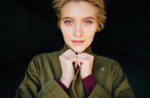 A person with light-colored eyes and blonde hair looks directly at the camera, wearing a green jacket and holding the collar with both hands. The background is dark, highlighting their face.