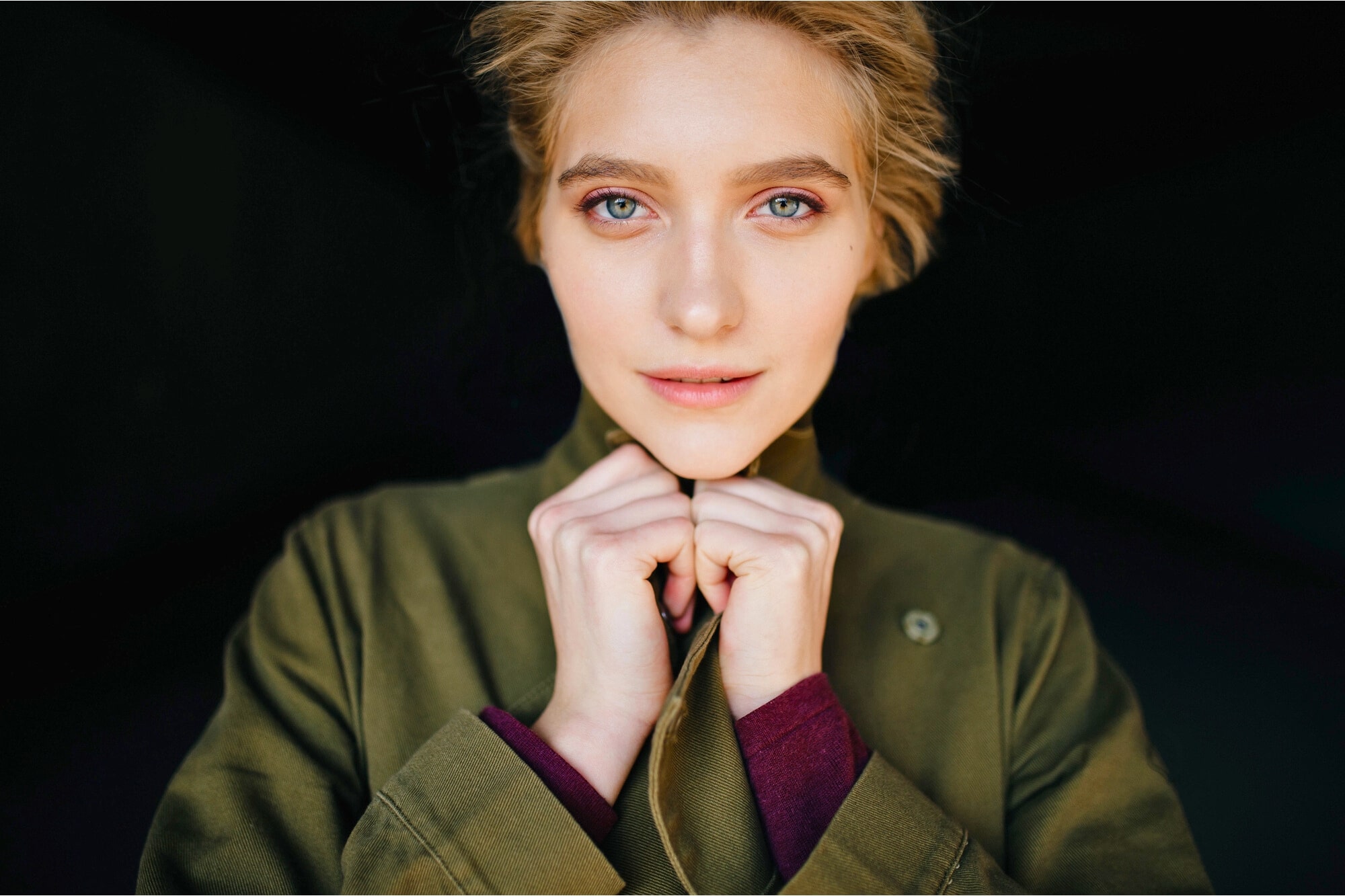 A person with light-colored eyes and blonde hair looks directly at the camera, wearing a green jacket and holding the collar with both hands. The background is dark, highlighting their face.