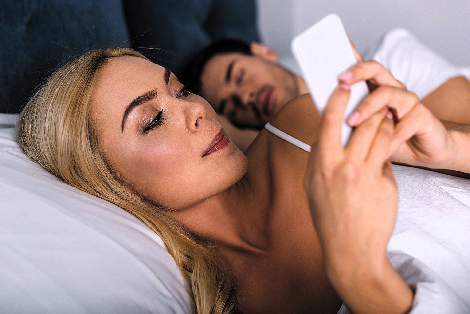 A woman lying in bed and using her smartphone, with a man sleeping beside her. She has long blonde hair and is focused on her phone. The bed has white sheets, and they are in a relaxed setting.